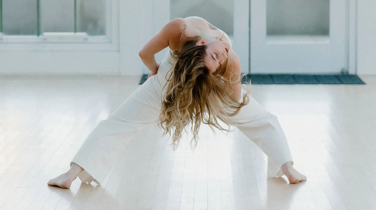 Молодая танцовщица в белом одеянии отклоняется назад к зрителю, ее волосы стекают к полу.