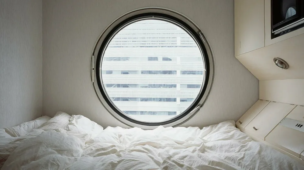 Interior of Nakagin Capsule Tower pod with round window and white bedding.