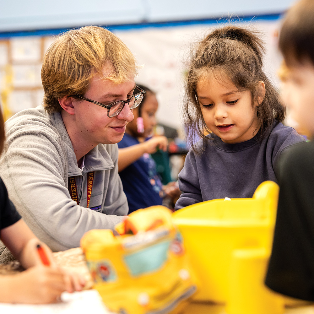 Adult volunteer assists young children with hands-on learning activity at classroom table surrounded by colorful bins and supplies materials.