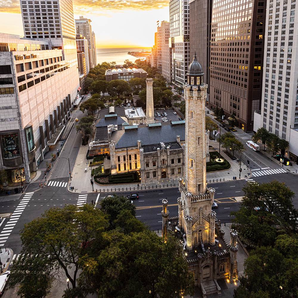  Aerial view of Chicago Water Tower surrounded by downtown skyscrapers at sunset, with tree-lined streets and historic masonry architecture.