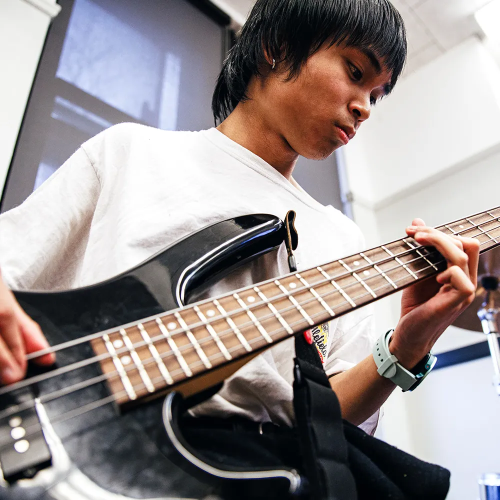 Student plays electric bass guitar during school music class, focusing on fretboard while sunlight enters through classroom windows nearby.