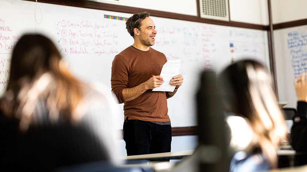  Instructor stands at front of classroom holding papers and marker, speaking to seated learners with whiteboard notes behind visible.