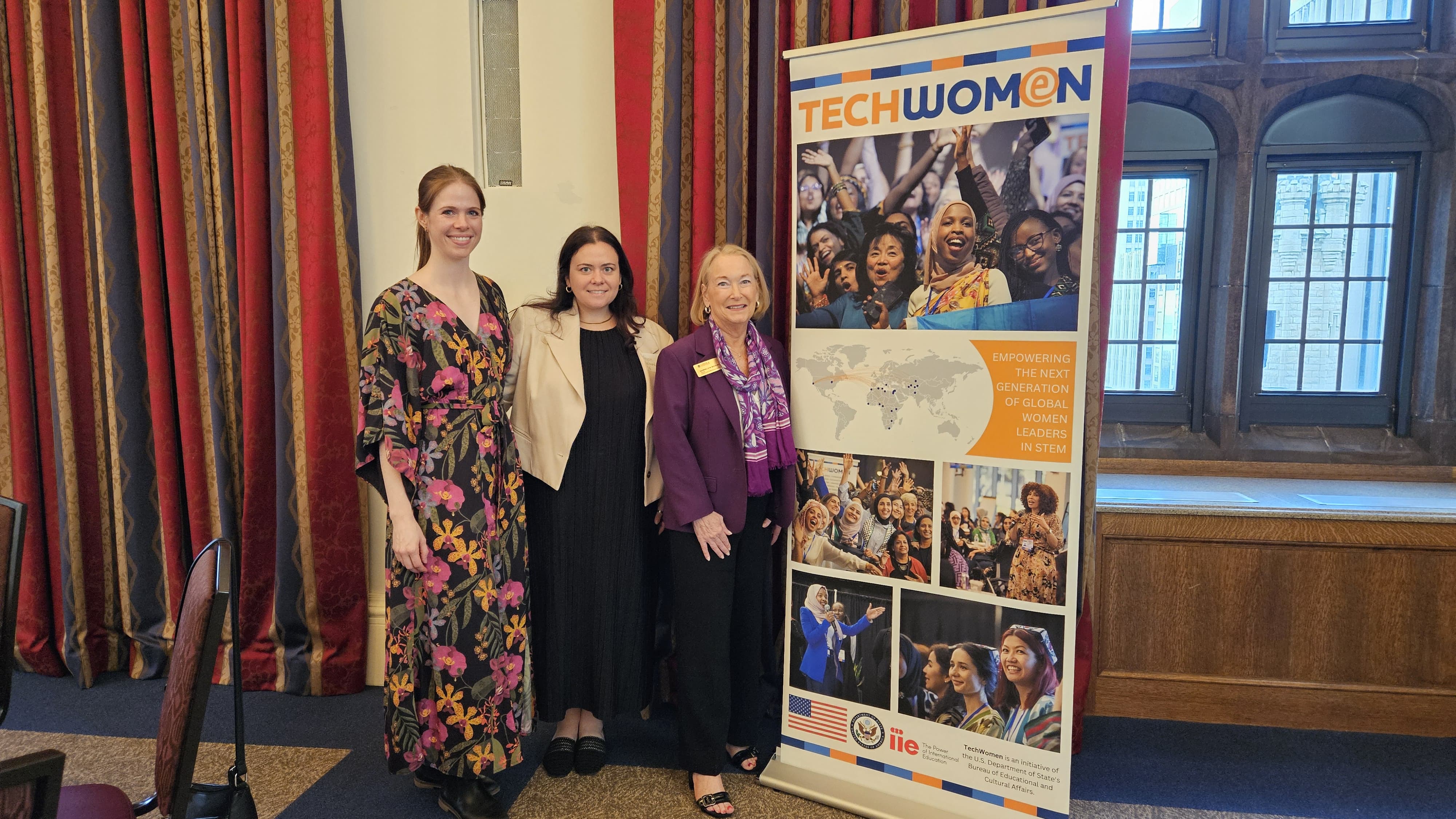 Three women stand in front of a colorful curtain next to a Tech Women banner