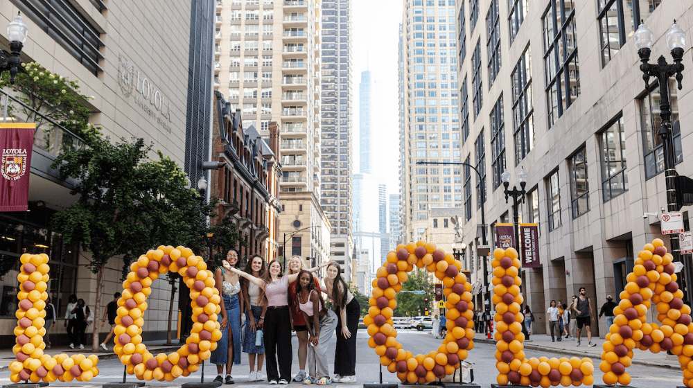 Students pose in downtown Chicago behind maroon and gold balloon letters spelling “LOYOLA,” with skyscrapers in the background.