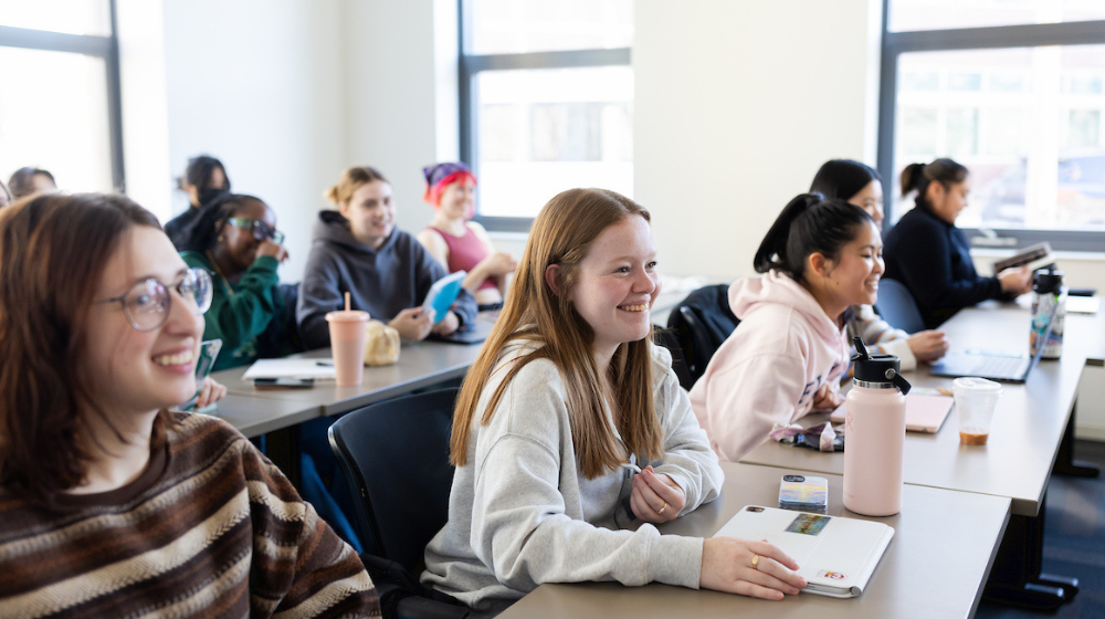 Students sit in a bright classroom, smiling and engaged during a lesson.