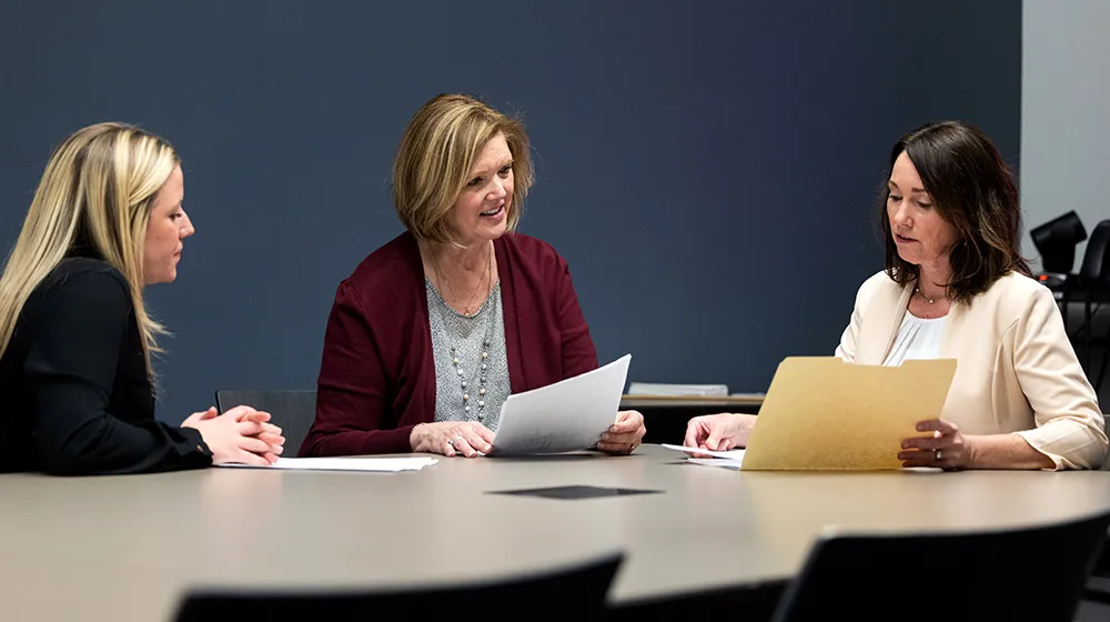 Three people review printed documents together at a conference table in a modern office meeting room with blue wall backdrop.
