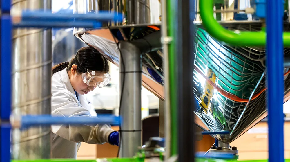 A laboratory worker wearing protective gear operates large stainless‑steel processing equipment surrounded by pipes and machinery