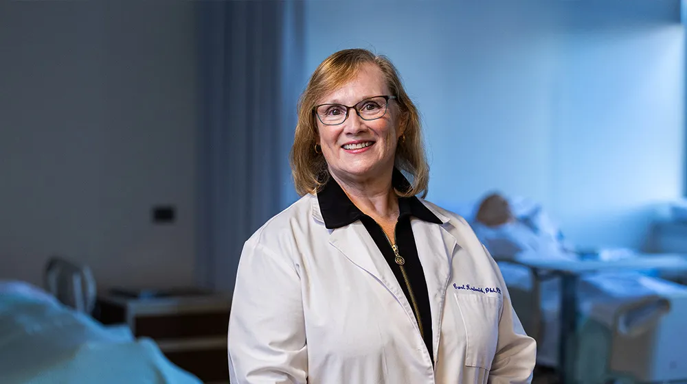 A healthcare professional in a white lab coat stands in a training room with hospital beds and medical equipment behind.