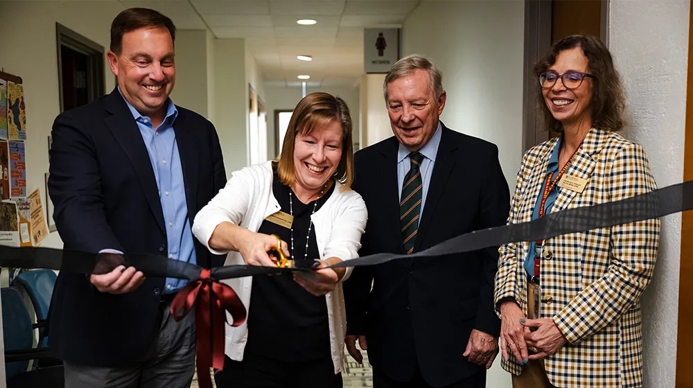  Four people participate in a ribbon cutting ceremony inside a hallway, holding oversized scissors and black ribbon during opening event.