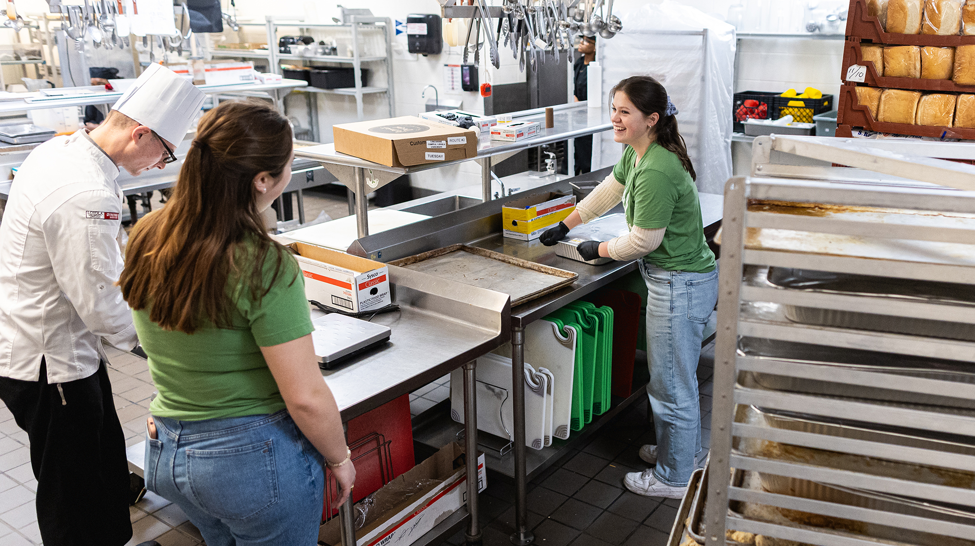 Two young woman students, dressed in green t-shirts and jeans, converse in a professional kitchen setting.