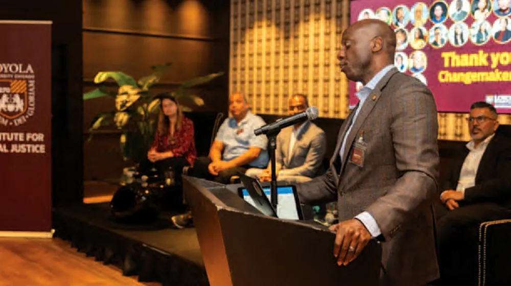 Man speaks at podium into microphone during panel event while attendees sit behind him on stage in formal venue.