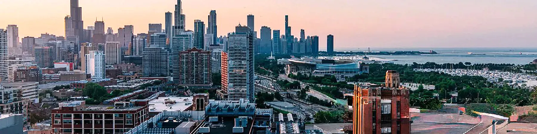 Chicago skyline at dusk shows dense high-rises, segments of winding highways. Lake Michigan stretches beyond rooftops under a soft, light coral sky.