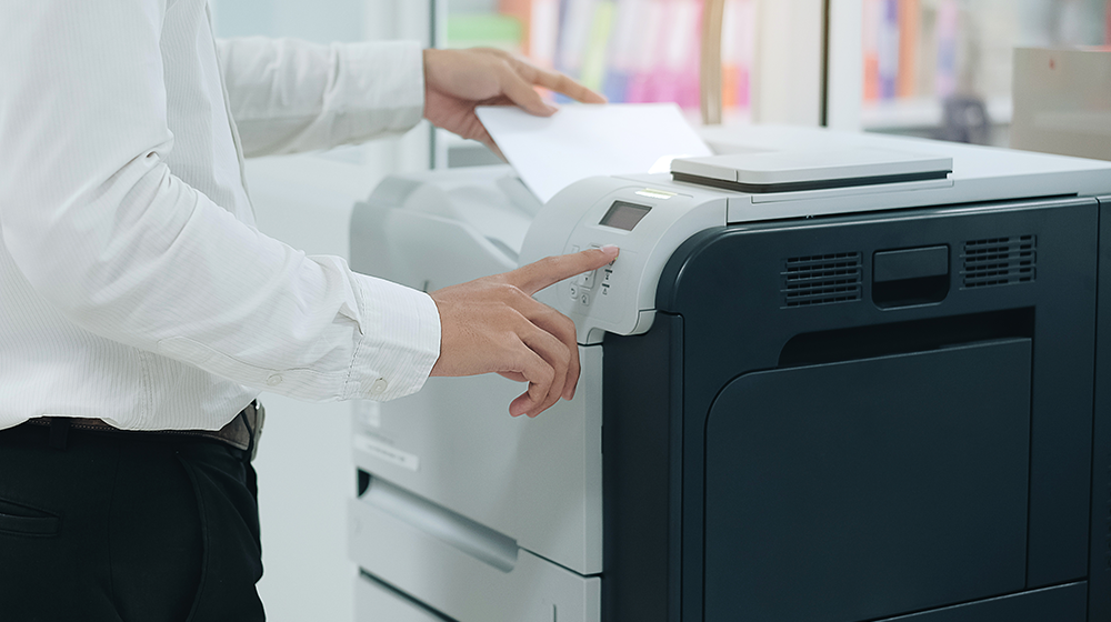 An individual stands in front of a computer lab printer retrieving their document as it finishes printing out.