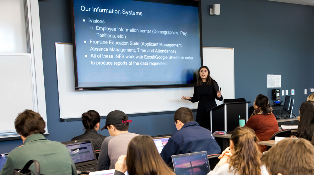 An instructor teaching with classroom technology and student using laptops.