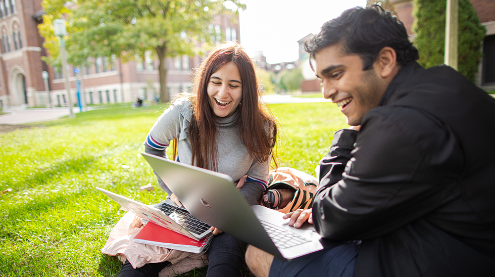 Two students seated on the grass in the West Quad near Damen Hall enjoy working together while studying with laptops and textbooks present.