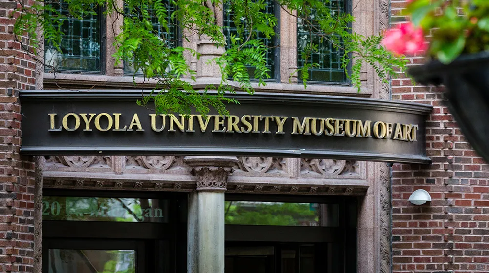 A group of brass serif letters spell out Loyola University Museum of Art on a dark brown background above the entrance to the museum. A group of brass serif letters spell out Loyola University Museum of Art on a dark brown background above the entrance to the museum.