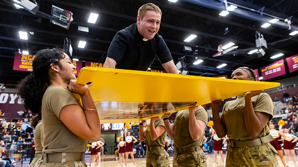 여섯 명의 ROTC 학생들이 보드를 들고 있고, 그 위에 예수회 신부가 푸쉬업을 하고 있는 모습
