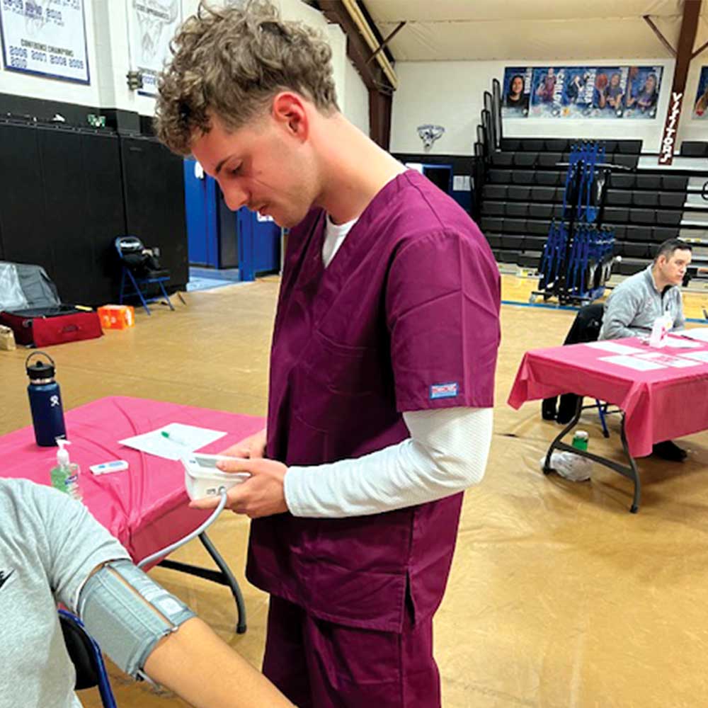 Человек в бордовых медицинских халатах измеряет артериальное давление другого человека, сидящего за столом во время мероприятия по скринингу здоровья.