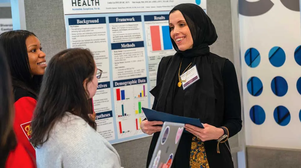 Человек в черном платке и черном костюме с золотым узором держит папку, представляя исследование на постерной сессии.