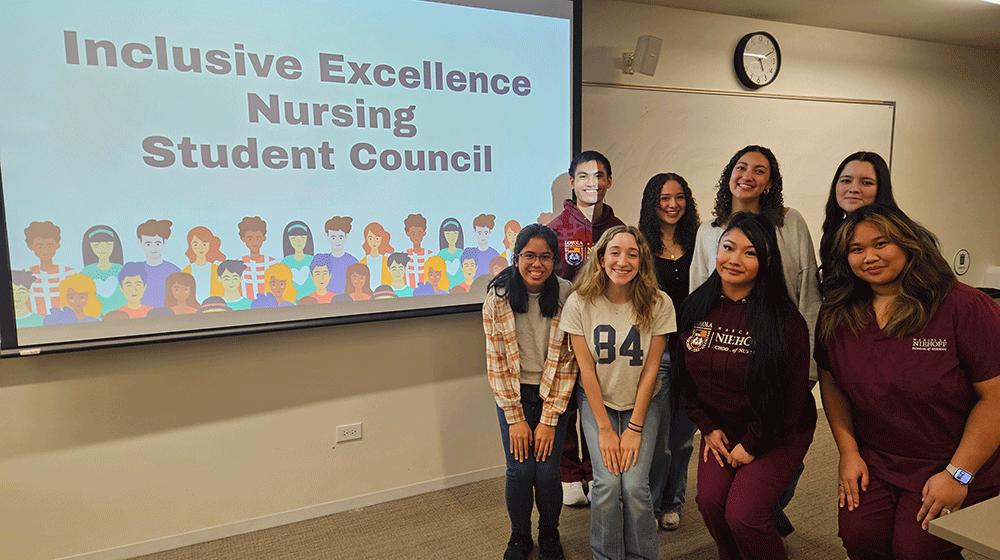Eight students, several in maroon scrubs, stand in a classroom next to a screen that says 