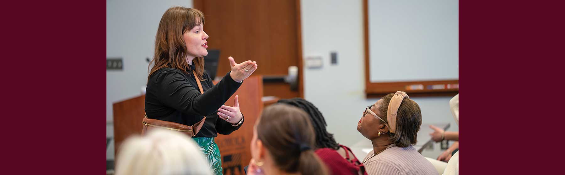 Person standing and gesturing while speaking to seated attendees in a classroom setting with a wooden podium and whiteboard visible.