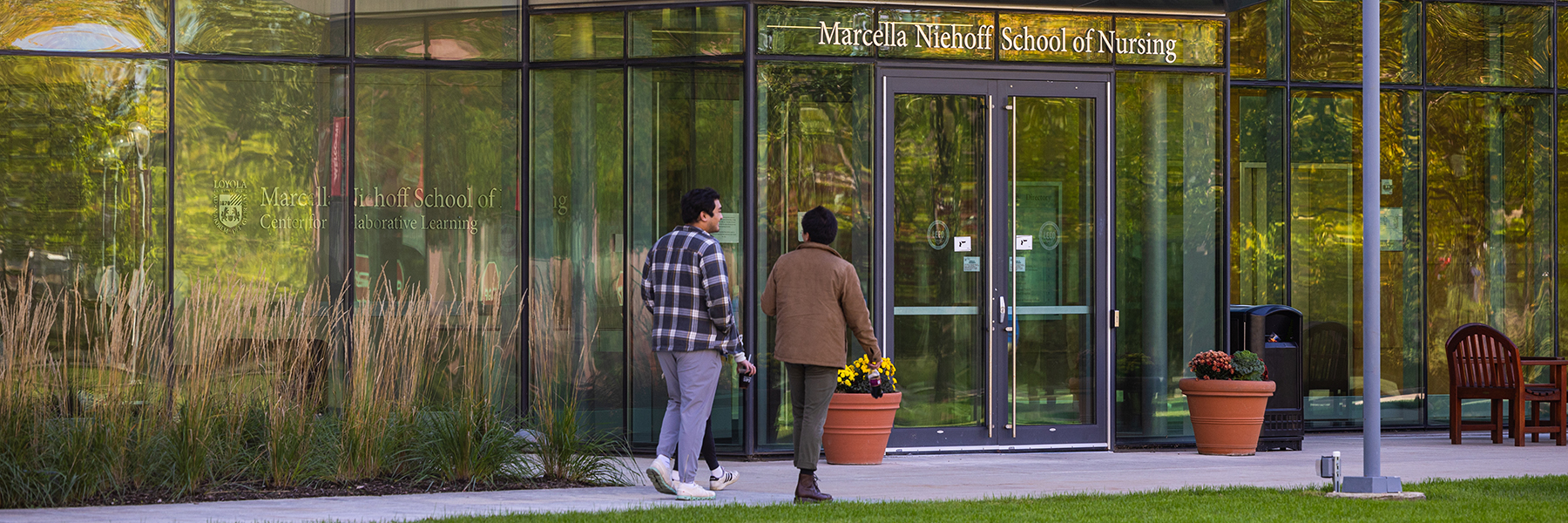 Two people walk outside HSC in front of the Nursing building on a sunny day, with the Marcella Niehoff School of Nursing's sign visible