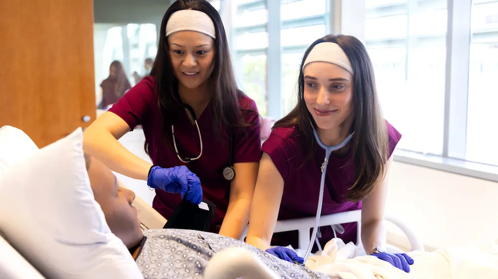 Two nursing students stand at the bedside tending to a clinical patient. Two nursing students stand at the bedside tending to a clinical patient.