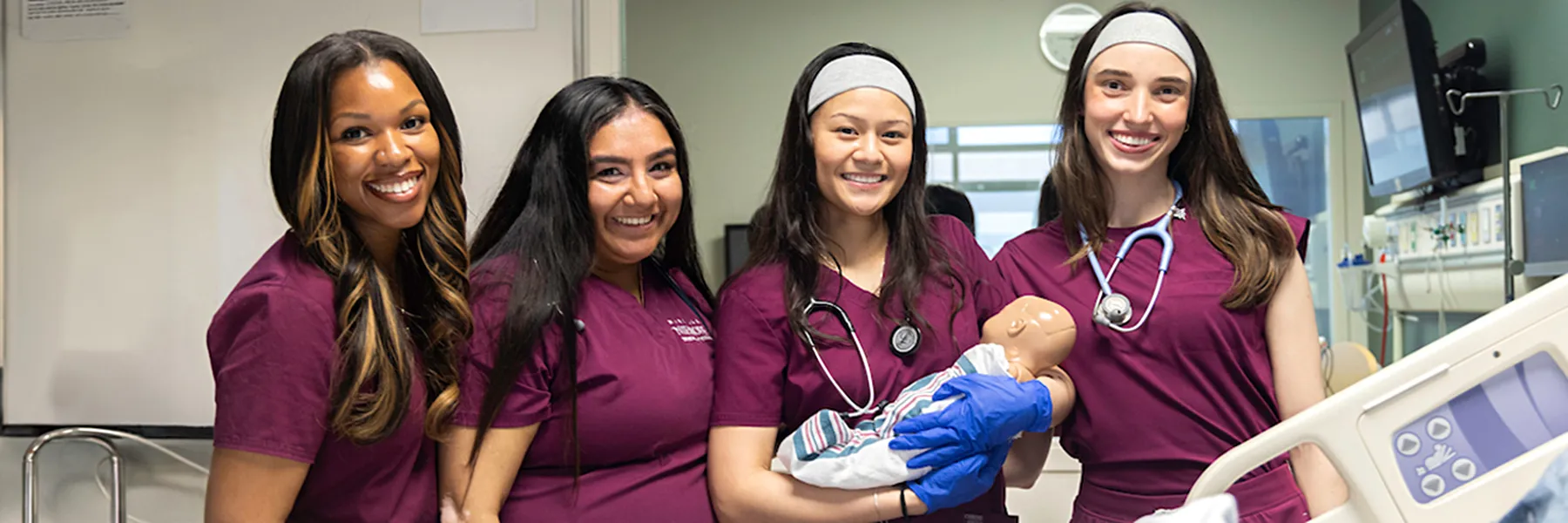 Four nursing students dressed in maroon Loyola scrubs stand side by side at a patient simulation station.