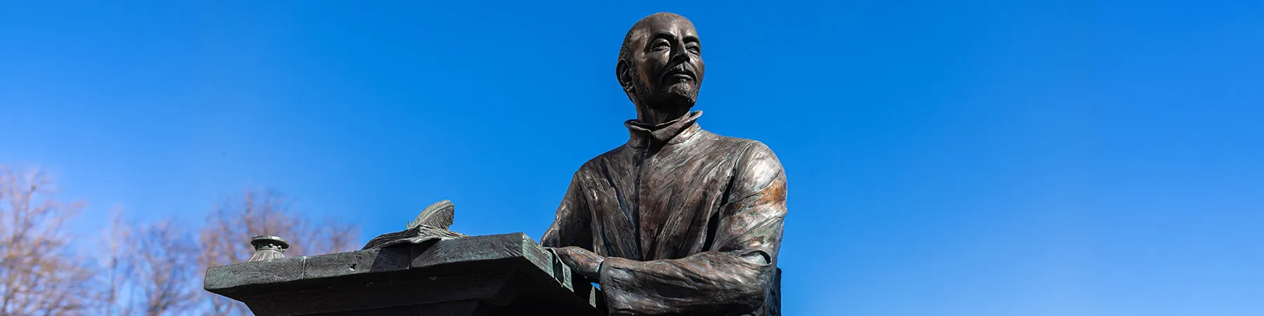 An outdoor, bronze, statue of St. Ignatius writing at a wooden desk with a quill pen in his hand. He looks to the side against a deep blue sky.