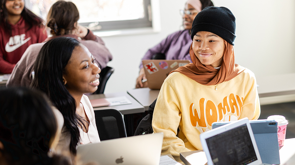 A smiling young African American girl wearing a dark beanie and a light yellow sweatshirt sits in a classroom interacting with other students near her