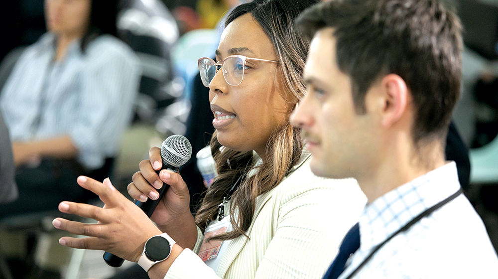 A young African American woman wearing glasses and a white coat holds a microphone and speaks to an unseen crowd around her.