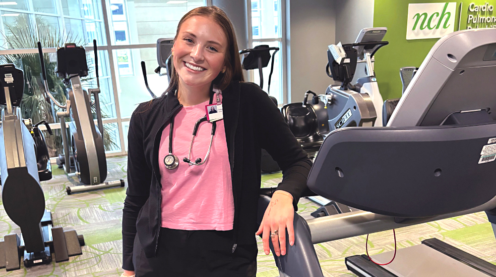 A young caucasian woman wearing a dark sweater over a pink shirt stands near a treadmill with her left elbow on one of the treadmill supports.
