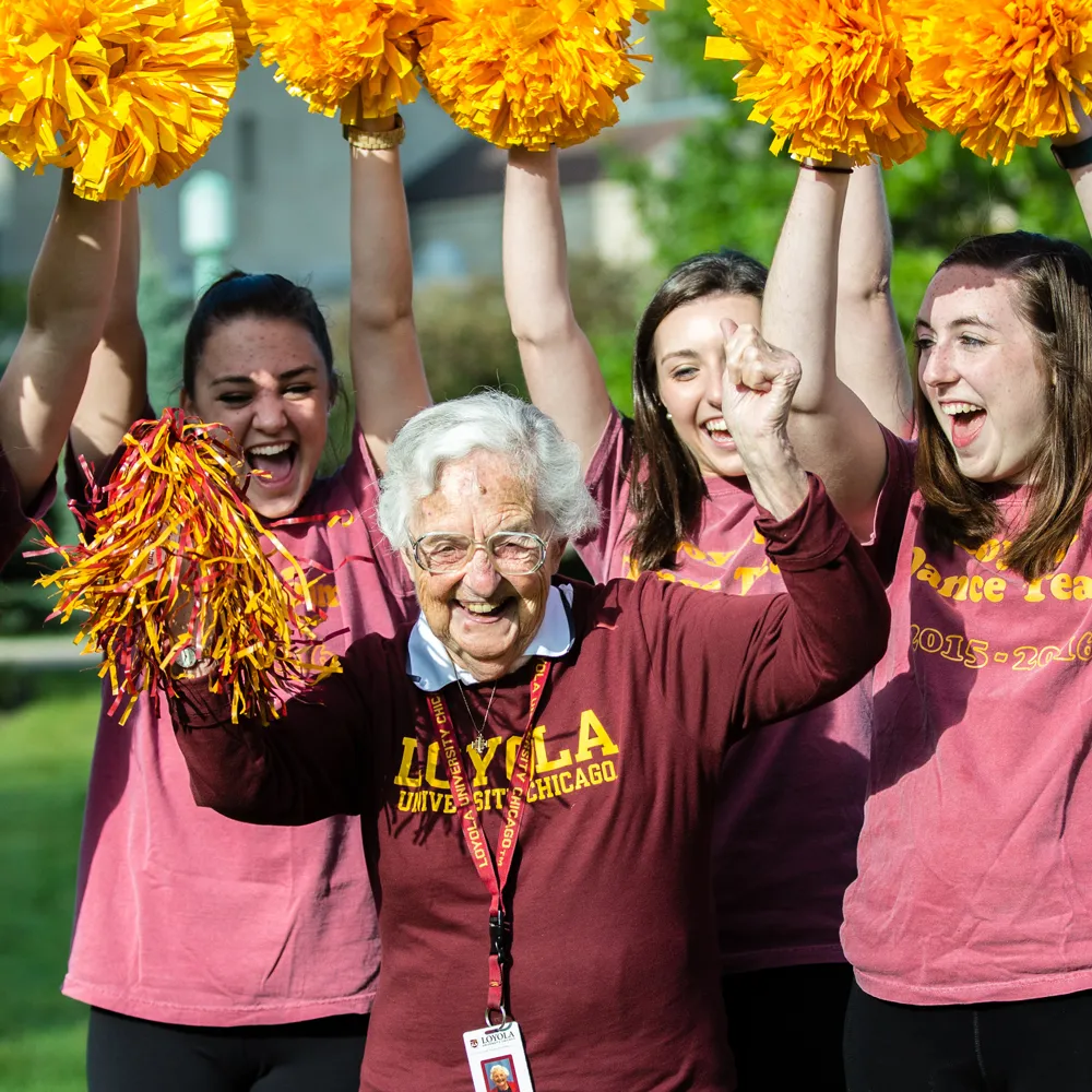Sister Jean holding maroon and gold pom poms, cheers with three cheerleaders. They all wear maroon sweatshirts or t-shirts tops.