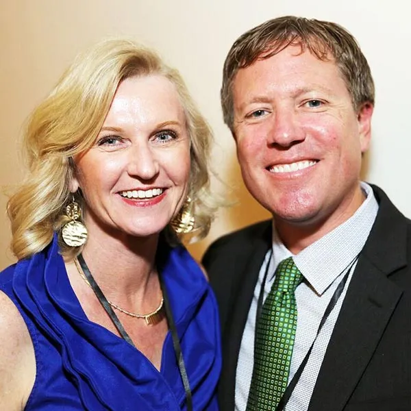 A man and a woman smile facing the camera against a beige background wall. The woman is wearing a blue shirt and the man is wearing a suit and tie.