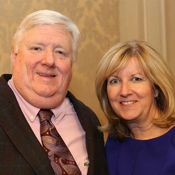 A man and a woman smile facing the camera against a beige background wall.