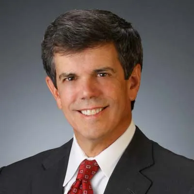 A man smiles for a portrait against a grey background. He is wearing a dark grey suit, white shirt, and a red tie.