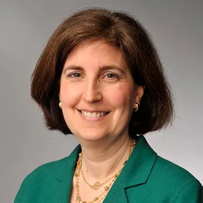 A woman in a green suit smiles while facing the camera against a grey background.
