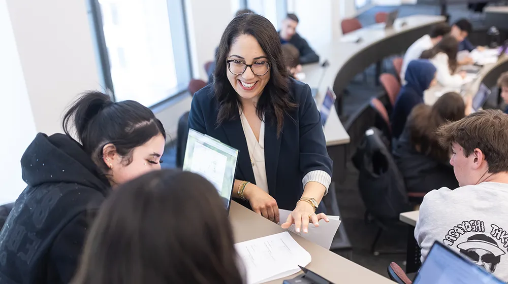 Irem Segal Orgit smailes as she interacts with students in her classroom. Several other students in the classroom can also be seen behind her.