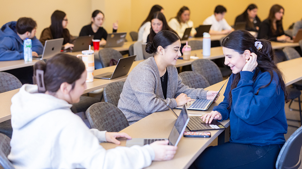Students interact while sitting in small groups of three or four. One student in the foreground is wearing a bright blue sweatshirt.