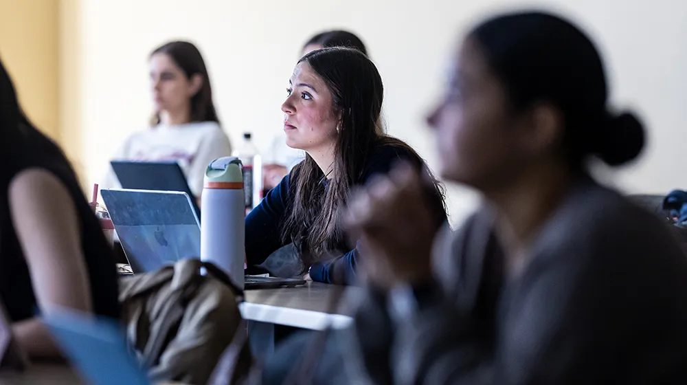 An attentive student sits amongst several students listening to a presentation. The low lit room is slightly darkened.