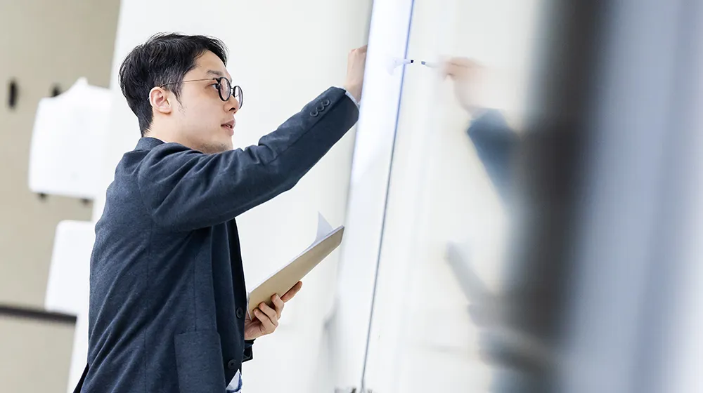 Yuxiao Rain Luo holds a notebook and writes on a whiteboard in a classroom.