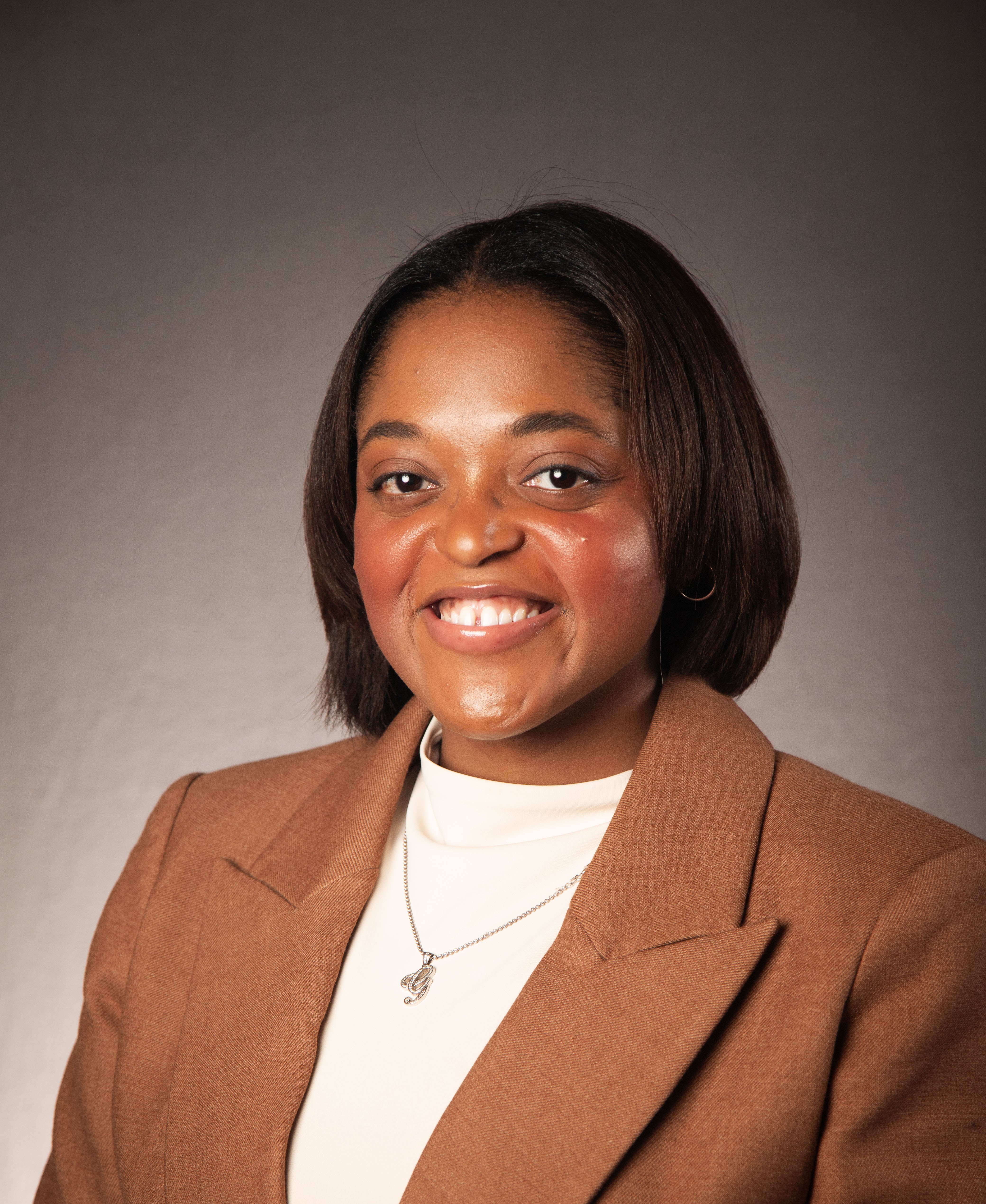 african american female wearing a brown suit jacket with a cream blouse and silver necklace with the letter G sitting in front of a gray background african american female wearing a brown suit jacket with a cream blouse and silver necklace with the letter G sitting in front of a gray background