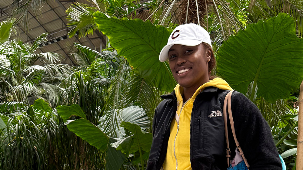 A person stands among large tropical plants in a lush indoor garden, wearing casual outdoor clothing and carrying a bag.