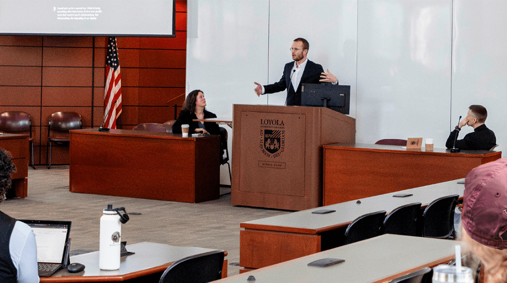 A speaker stands at a podium addressing an audience in a university classroom while panelists listen and presentation slides display.