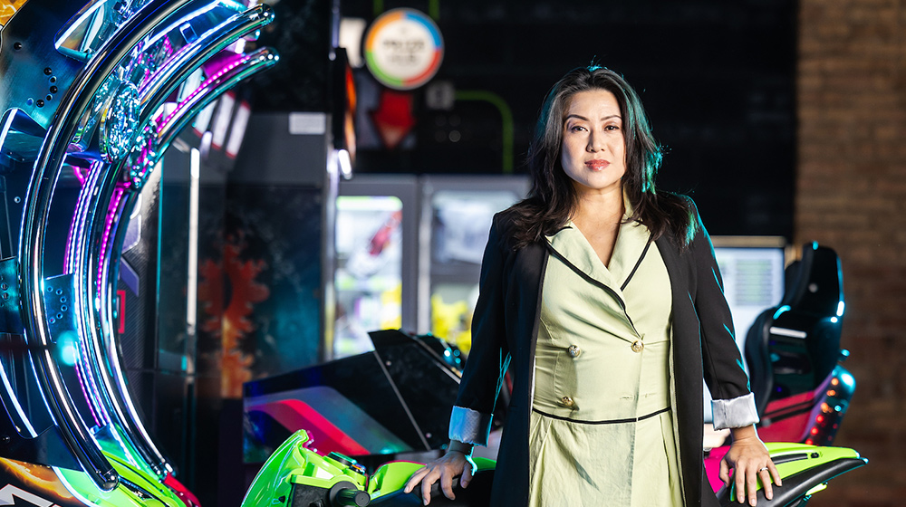 A person stands between brightly lit arcade racing games, wearing a light dress and dark jacket in a colorful gaming environment.