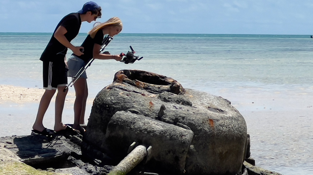 Two people set up a camera beside a rusted metal structure on a sunny beach overlooking calm turquoise ocean water.