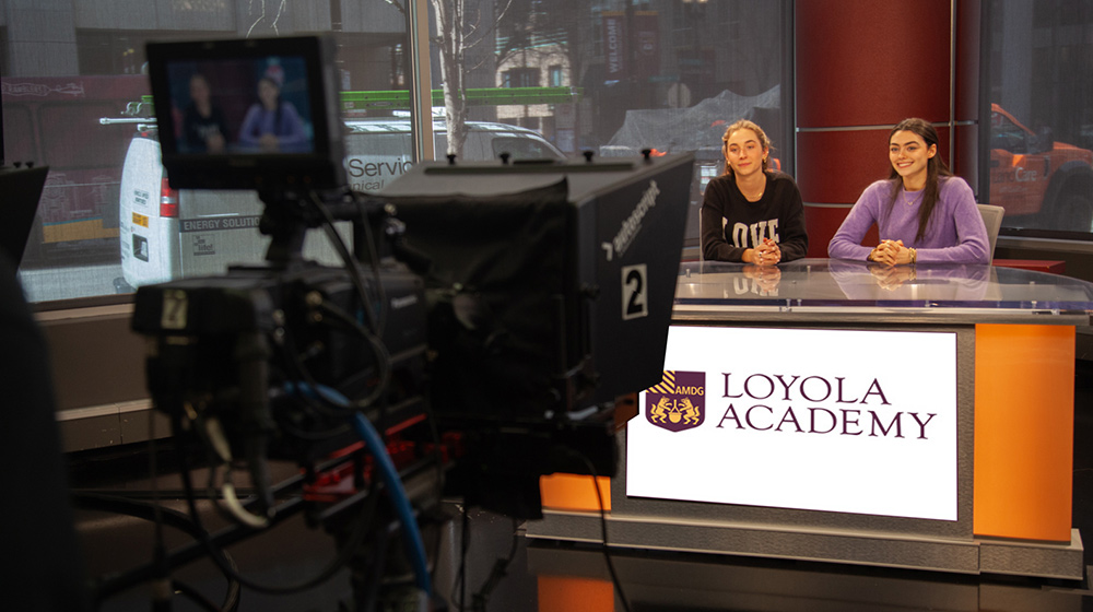 A television studio scene with two people seated at a news desk while a large broadcast camera records the segment.