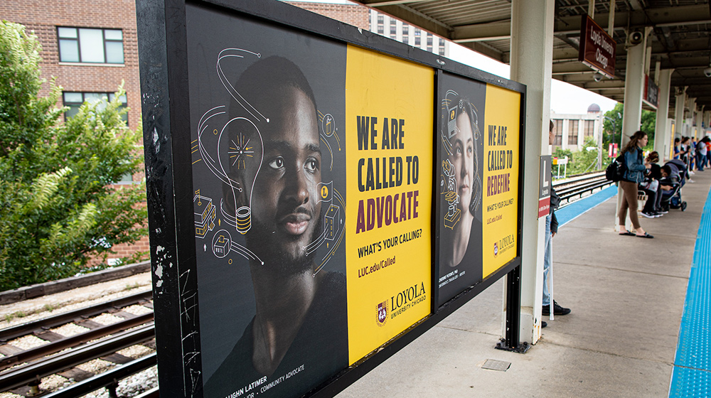 Advertising posters promoting advocacy and service for a university are displayed along a busy outdoor train platform with commuters waiting.