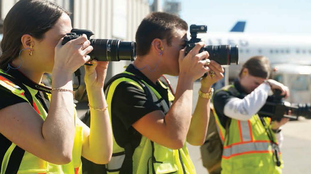 Photographers wearing safety vests take pictures on an airport tarmac, aiming their cameras toward aircraft and activity in the distance.