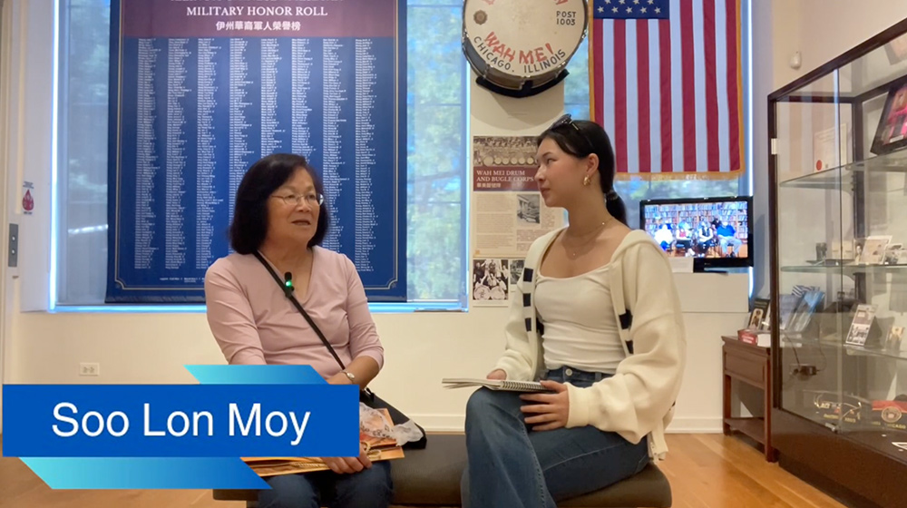 Two people sit together in a museum space conducting an interview, surrounded by historical displays, memorial plaques, and an American flag.